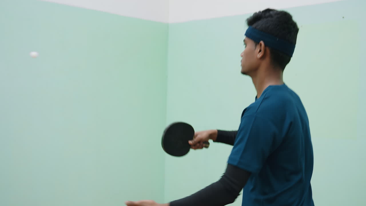 man in blue shirt and headband practicing tennis against wall indoors, holding paddle and focusing on hitting ball, showing determination, training effort, and concentration during practice session