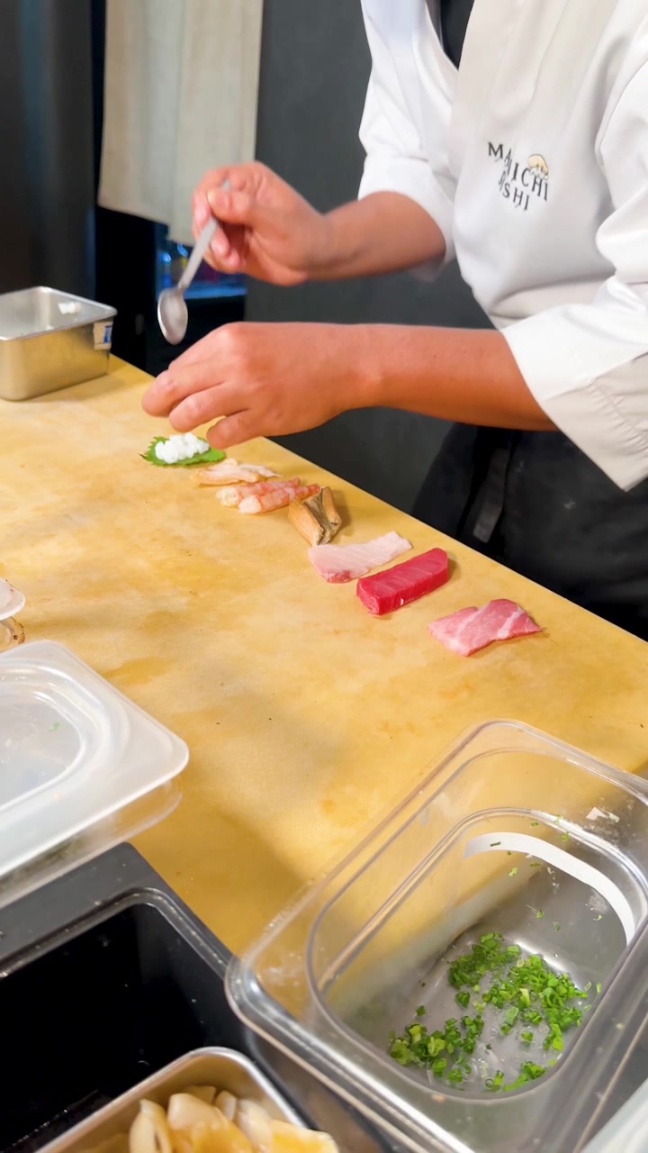 明るい頭上照明の下、木製のカウンターで寿司飯と魚を握るプロの寿司職人。