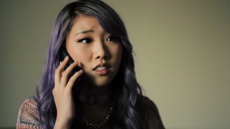 a young woman with purple hair engaged in a serious phone conversation, displaying concern and emotion in a subdued background setting
