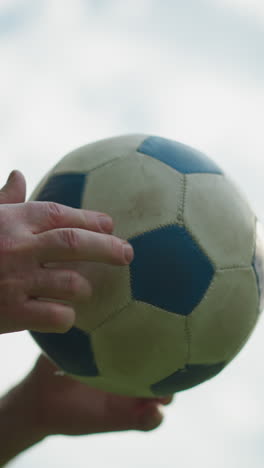 a side view of an elderly man gently rolling a soccer ball in his hand, with a cloudy sky seen