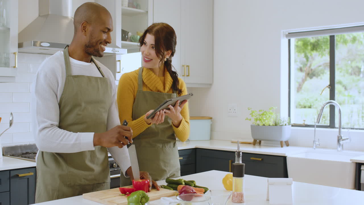 Diverse multiracial couple cooking together in kitchen, using tablet for recipe guidance, at home