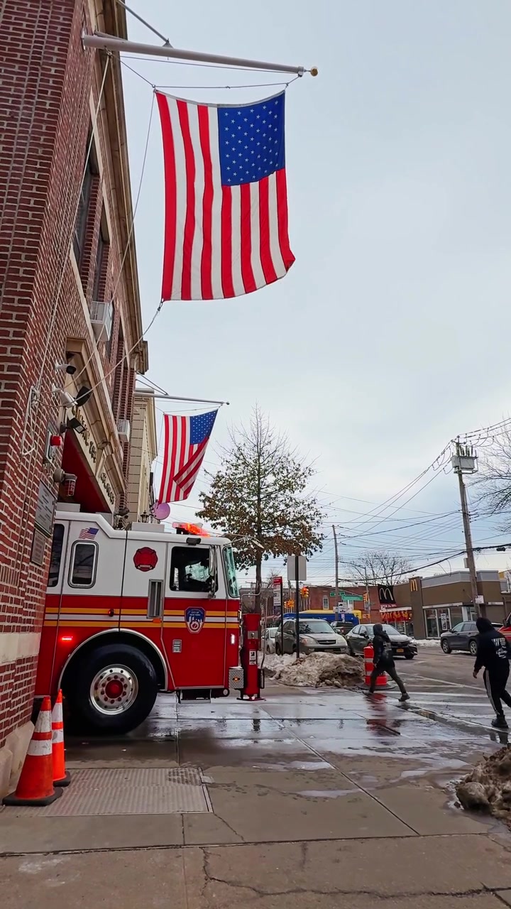 New York, USA, 25 gennaio 2026: Camion dei pompieri rosso 153 parcheggiato davanti a una caserma dei pompieri in mattoni. Ampia inquadratura di un camion dei pompieri fuori da un tradizionale edificio di caserma dei pompieri in mattoni con bandiere americane che sventolano.