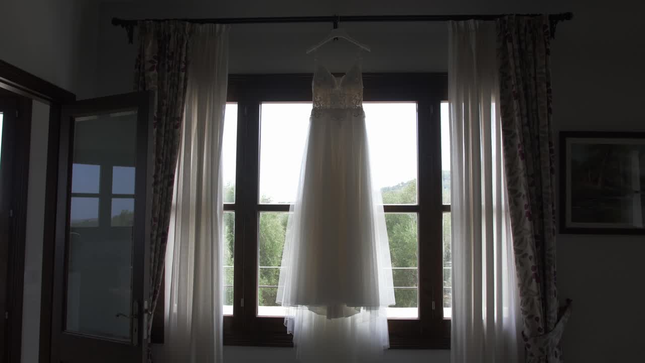 Abito da sposa appeso davanti a una finestra all'interno di una tradizionale finca di Maiorca con luce diurna soffusa e lento movimento di macchina.