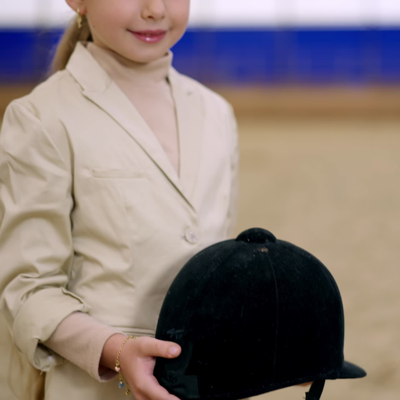 Beautiful young long-haired girl holding a black jockey helmet in her hands. Cute kid smiling to the camera standing still. Riding hall at backdrop in blur