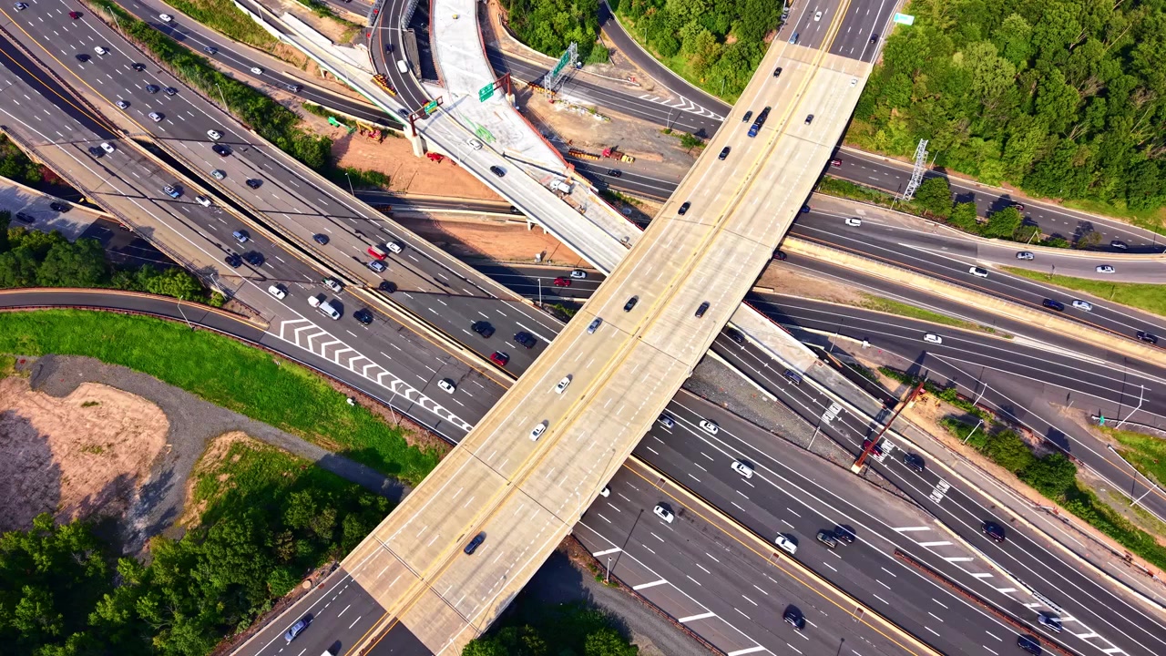 Overhead shot of X shaped overpass crossing multilane freeway. Drone perspective of construction zone, concrete ramps and moving traffic beside forest. Garden state parkway.