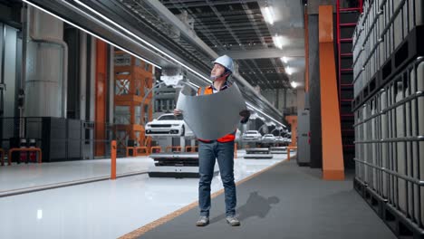 cuerpo lleno de ingeniero masculino asiático con casco de seguridad mirando el plano en sus manos y mirando a su alrededor mientras estaba de pie en la fábrica de automóviles ev