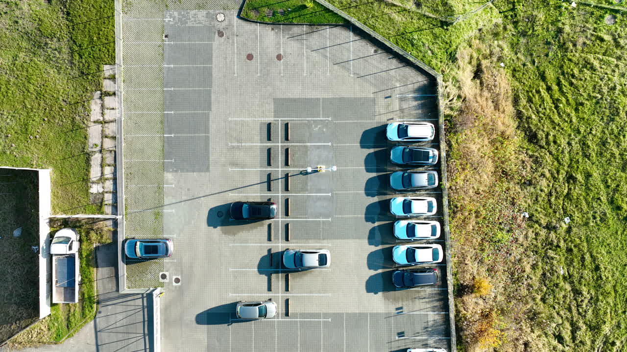 Oversiktsbilde ovenfra av et lite parkeringsområde med flere parkerte biler nær en industribygning og en gresskledd kant i sollys