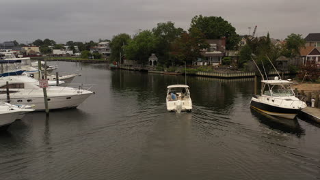 a low angle, aerial view of a fishing boat sailing through a canal behind houses on long island on a cloudy day