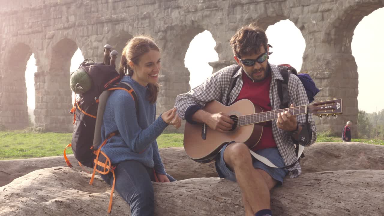 giovane coppia di backpackers turisti seduti su un tronco di legno suonando la chitarra cantando di fronte all'antico acquedotto romano rovine nel romantico parco degli acquedotti parco a roma all'alba sacco a pelo movimento lento