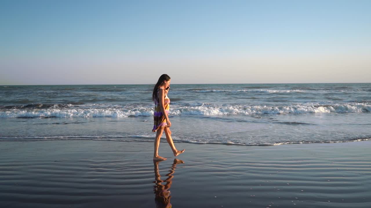 Female Portrait Walking Along Seashore While Talking On Her Smartphone. Static Shot