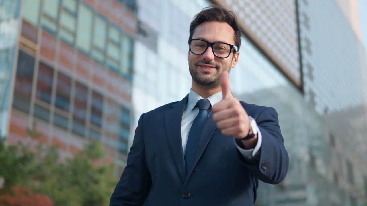 joven hombre de negocios barbudo vestido con traje mirando a la cámara mostrando el pulgar grande cerca de los edificios de la torre grande