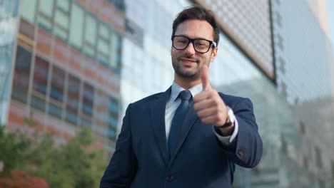 young bearded businessman dressed in suit looking to the camera showing big thumb up near large tower buildings