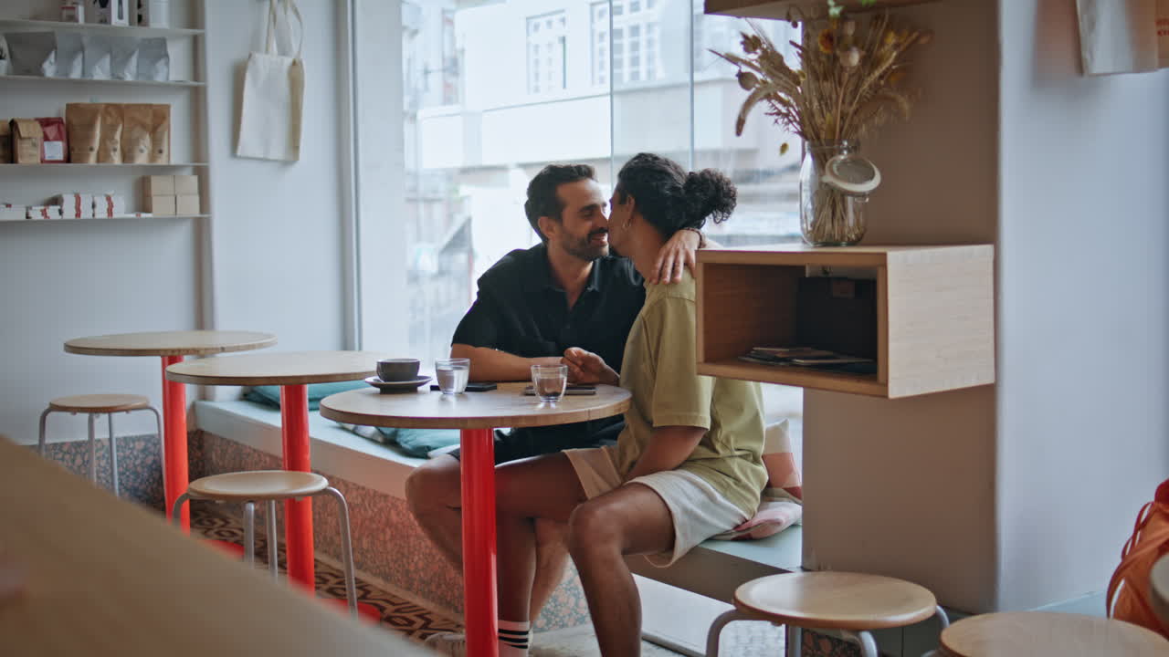 Coppia gay spensierata a un appuntamento al tavolo di un caffè. Due uomini multietnici si abbracciano e si baciano.