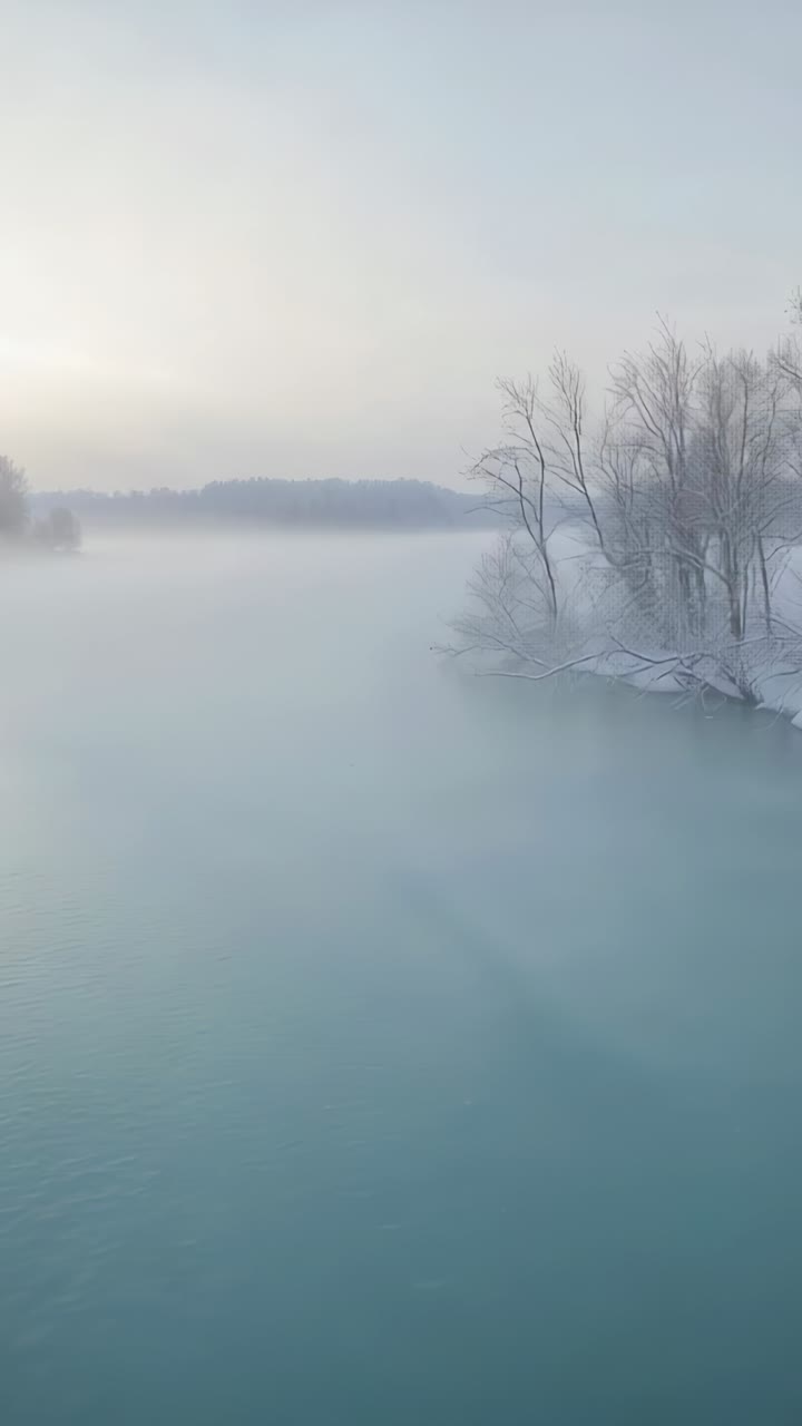 Vertical video: Mist drifting across teal river and snowy trees, cold air meeting warmer water