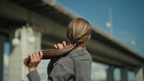 lady straightens her hair while looking away near a bridge, she adjusts the front part of her hair as she flaunts the length, with blurred electric poles and structures in the background