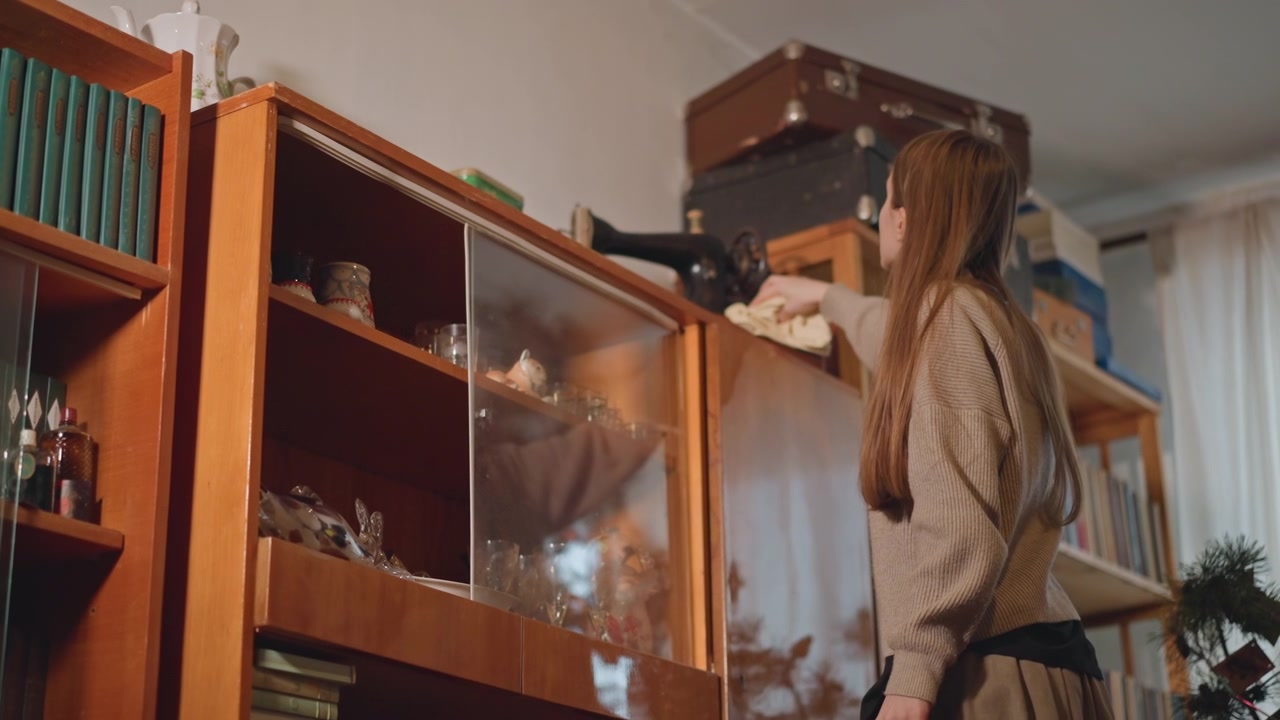 Caucasian woman reaches for box on top shelf in cozy library interior, braids fall over shoulder as she lifts dusty case from cabinet display, deliberate movement reveals curated collection of books