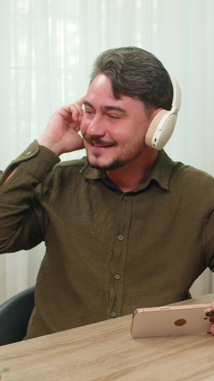 Middleaged man laughs listening funny audio smartphone headphones bright mood joyful vibe at table