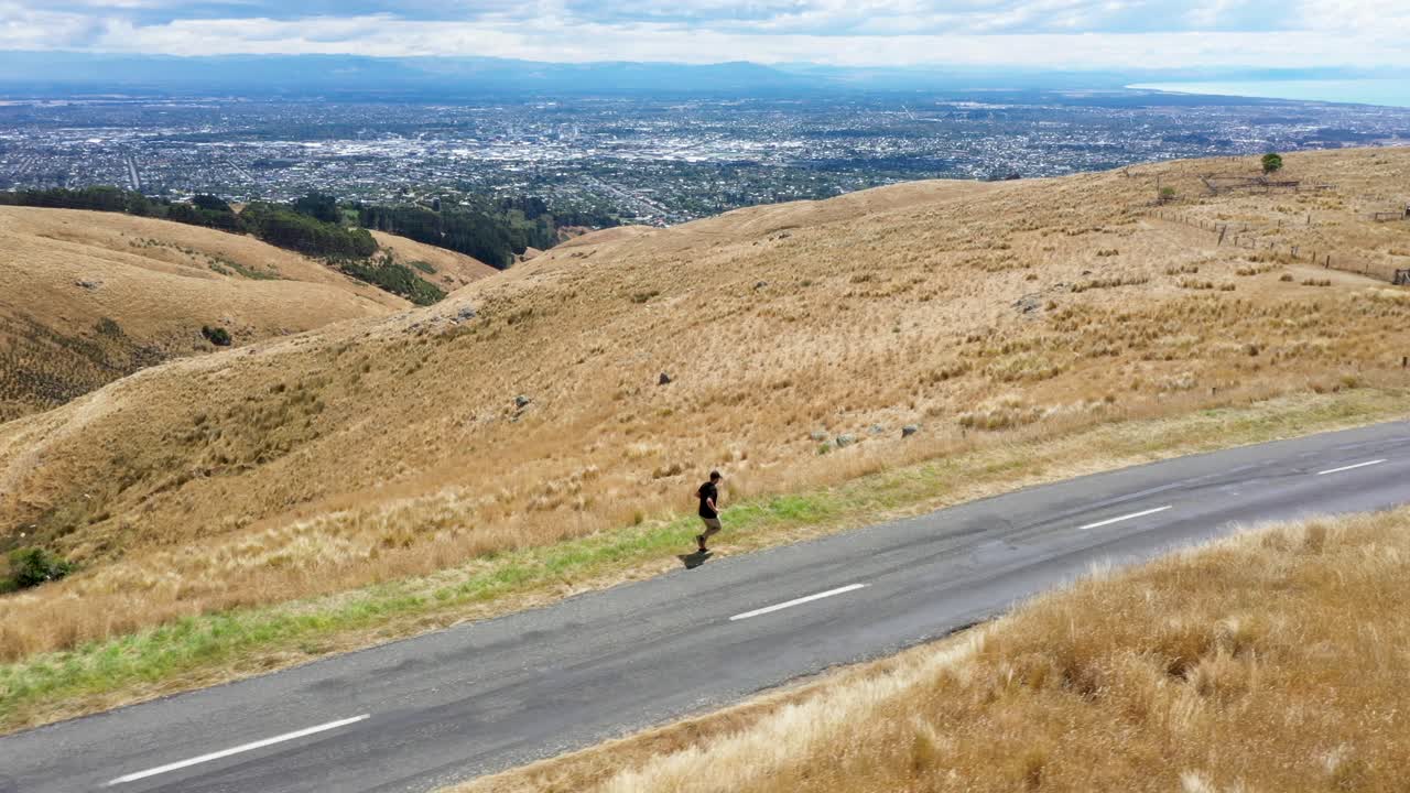 Follow shot of man running in hills next to road with city in background.