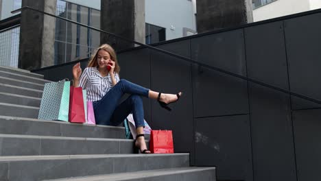 Girl-sitting-on-stairs-with-bags-talking-on-smartphone-about-sale-in-shopping-mall-in-Black-Friday