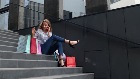 girl sitting on stairs with bags talking on smartphone about sale in shopping mall in black friday