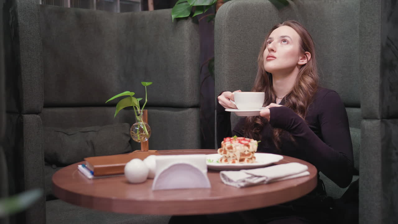 Young woman in dark outfit lifts white cup of latte to face with closed eyes, inhaling aroma while seated in green booth. Wooden table features waffle, napkin, ceramic decoration, and leafy plant