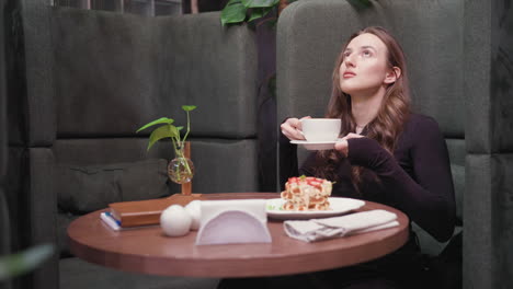 young woman in dark outfit lifts white cup of latte to face with closed eyes, inhaling aroma while seated in green booth. wooden table features waffle, napkin, ceramic decoration, and leafy plant