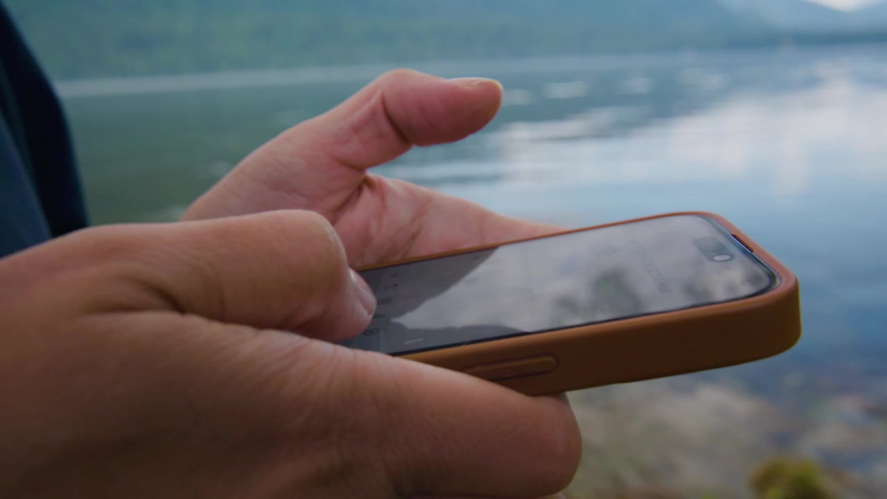 Primo piano di una mano che tiene uno smartphone vicino all'acqua. Le dita toccano delicatamente lo schermo. La persona appare calma e concentrata. Lo sfondo mostra un lago calmo con colline in lontananza. Ideale per viaggi, tecnologia.
