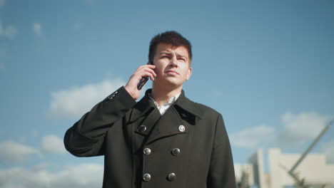 man holding phone outdoors, preparing to answer incoming call, dressed in black coat with metallic buttons, standing under bright blue sky with blurred background featuring greenery