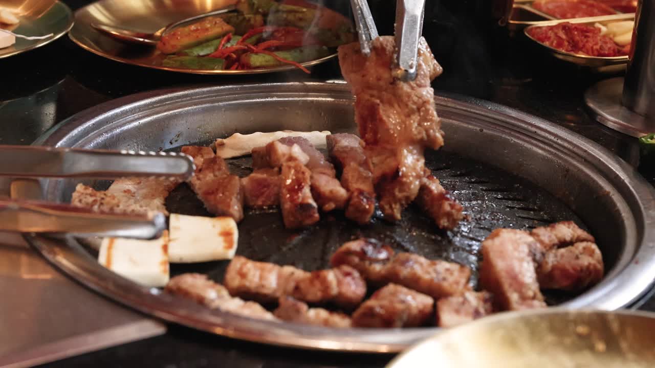 Hand uses tongs and scissors to cut pork belly on tabletop grill, warm restaurant lighting