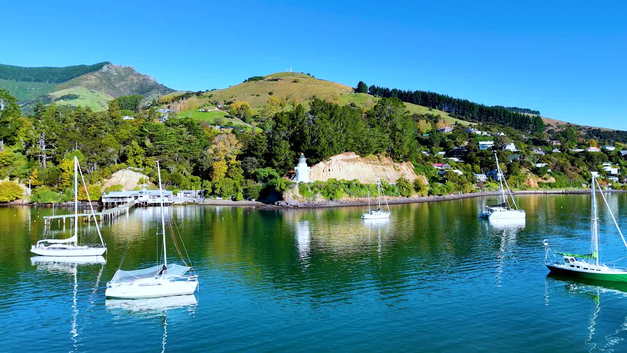 Aerial view of Akaroa Harbour showcasing boats, lush hills, and a lighthouse under clear blue skies