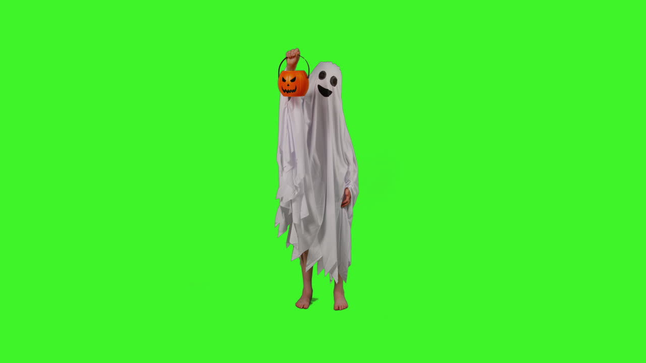 toma completa de un niño vestido con disfraces de fantasma haciendo trucos o tratamientos en halloween sosteniendo un cubo en forma de calabaza jack o'lantern contra la pantalla verde del estudio