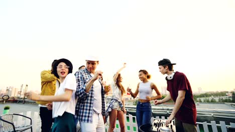 cheerful dj is working with mixing console and talking to young man dancing on rooftop at modern party. men and women are wearing trendy clothes and holding bottles.