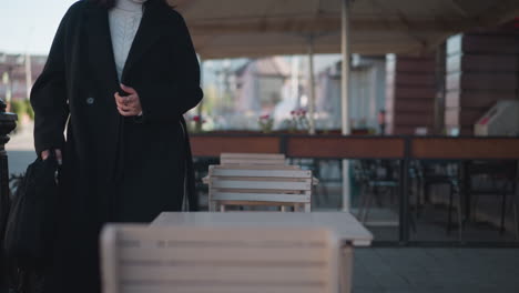 businesswoman in black coat and white turtleneck sits down at cafe table, placing her bag with an air of ease, background includes blurred cars, poles, and urban buildings under a shaded patio