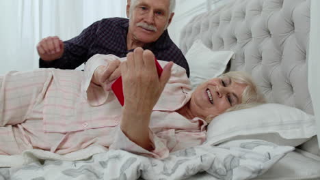 Senior-elderly-couple-wearing-pajamas-lying-on-bed-looking-on-mobile-phone-laughing-and-having-fun