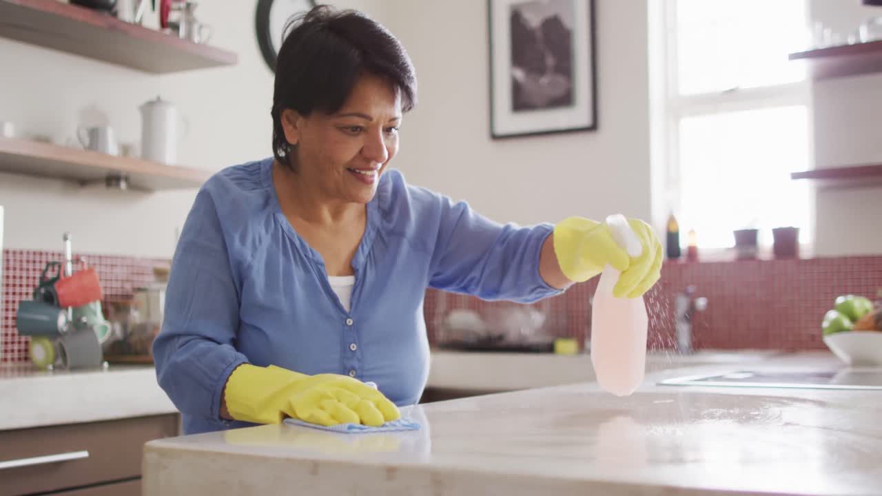 sonriente mujer biracial mayor con guantes y mesa de limpieza en la cocina sola