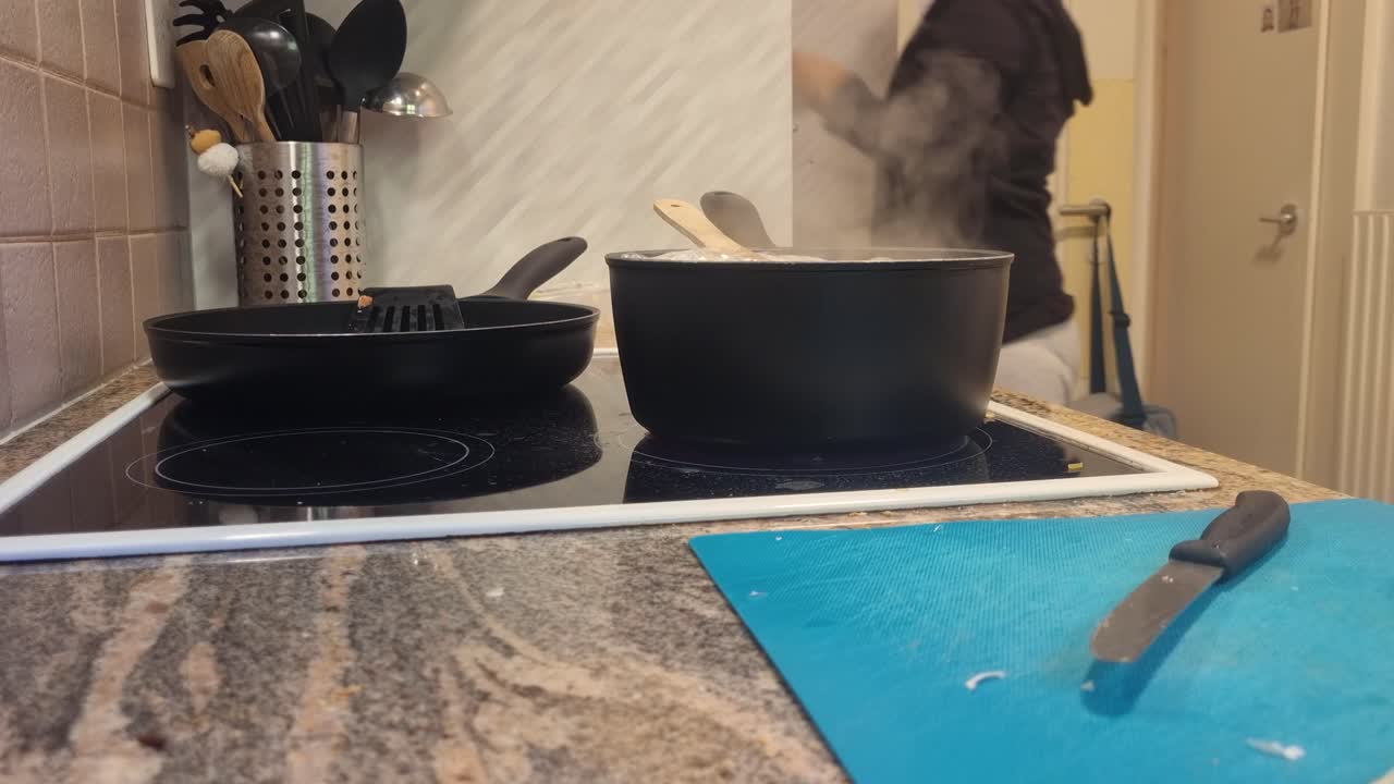 A woman cooks at home, stirring vegetables in hot water while removing crispy bacon cubes from a frying pan, Bern, Switzerland