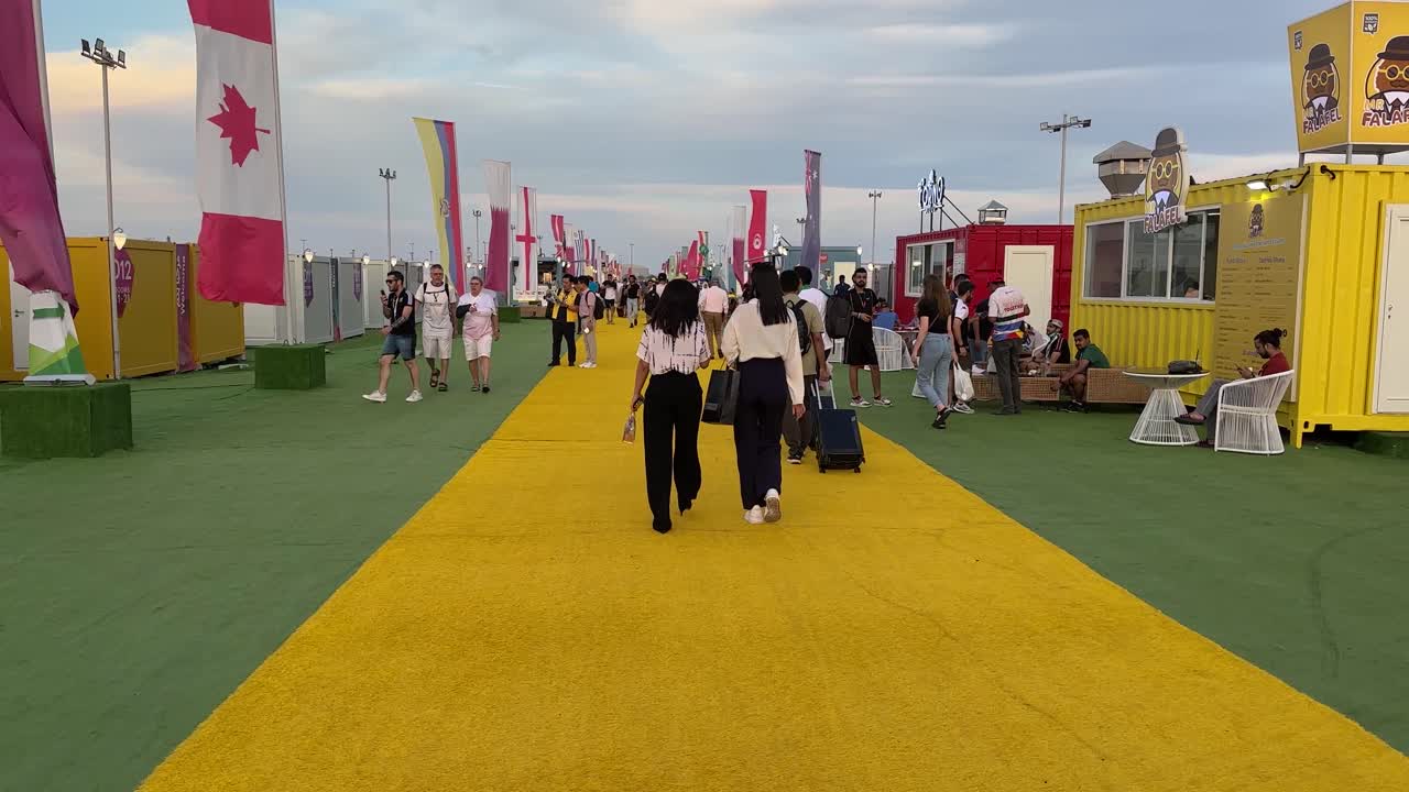 two women with dark black hairs white shirt and black trousers are walking in a yellow green floor in the fan village in Doha Qatar designed with flags and suits for accommodation in a colorful place