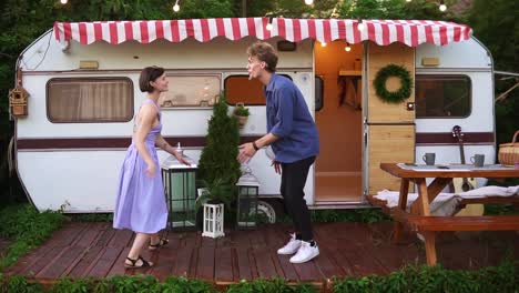 Stylish,-cheerful-couple-man-and-woman-smiling-while-dancing-together-near-house-on-wheels-outdoors.-Funky-dancing-in-front-the-trailer.-Retro-style-exterior.-Slow-motion.-Front-view