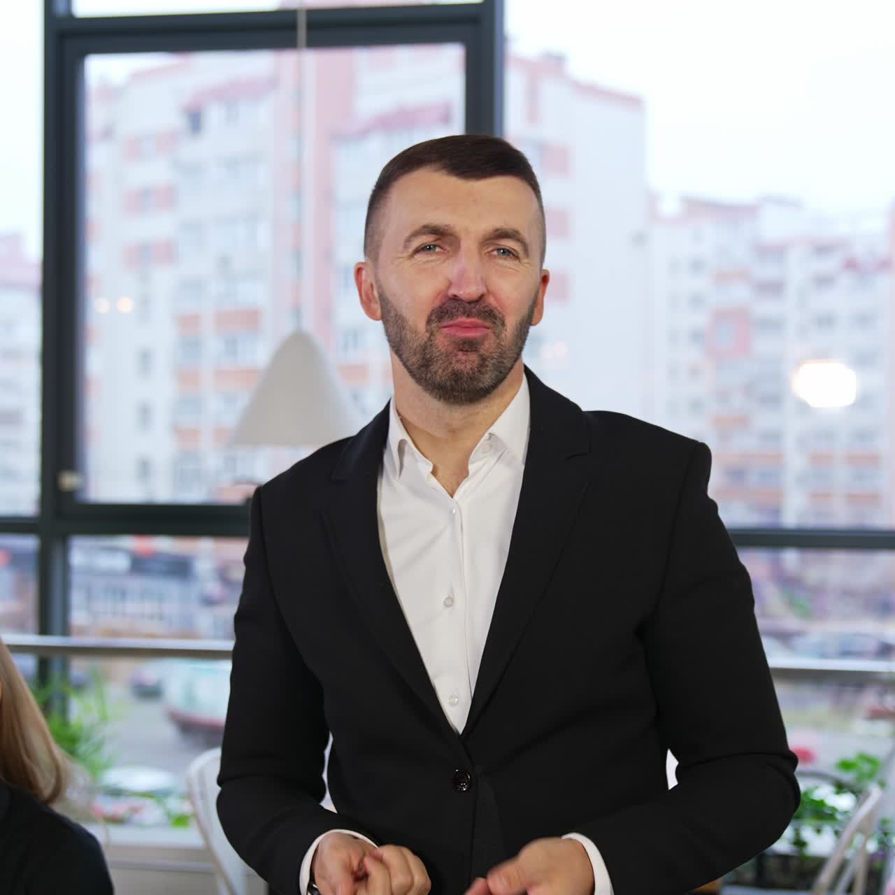 Bearded good-looking man speaking to the camera with a smile. Office workers sitting beside approvingly nod and discuss the speech