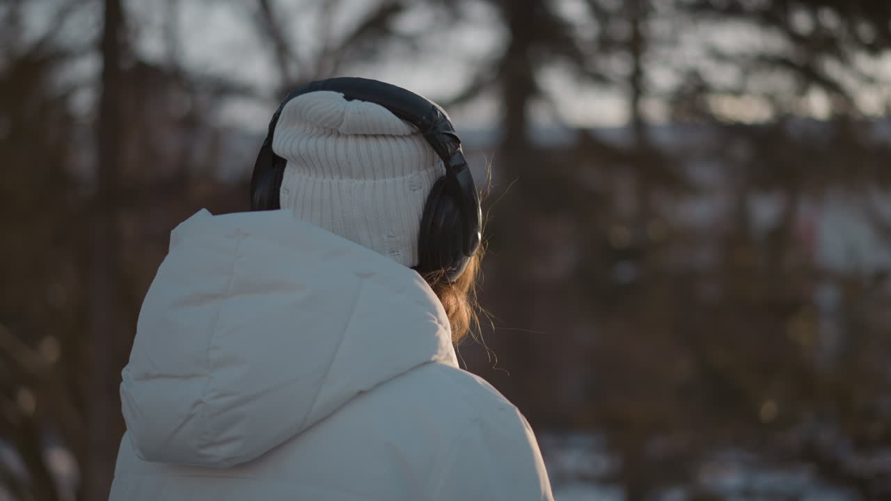 Side view of woman in white puffer jacket and beanie dancing under warm sunset light wearing headphones captured in flowing motion against blurred winter nature backdrop evoking serene energy