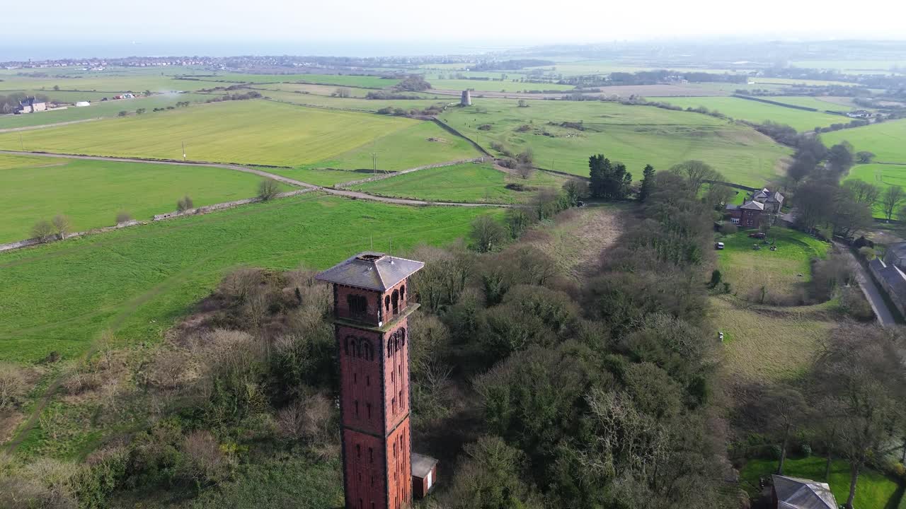 Cleadon Water Tower drone forward flight over historic brick landmark rising from woodland, with green fields, rural roads and scattered homes in Cleadon, South Tyneside, England