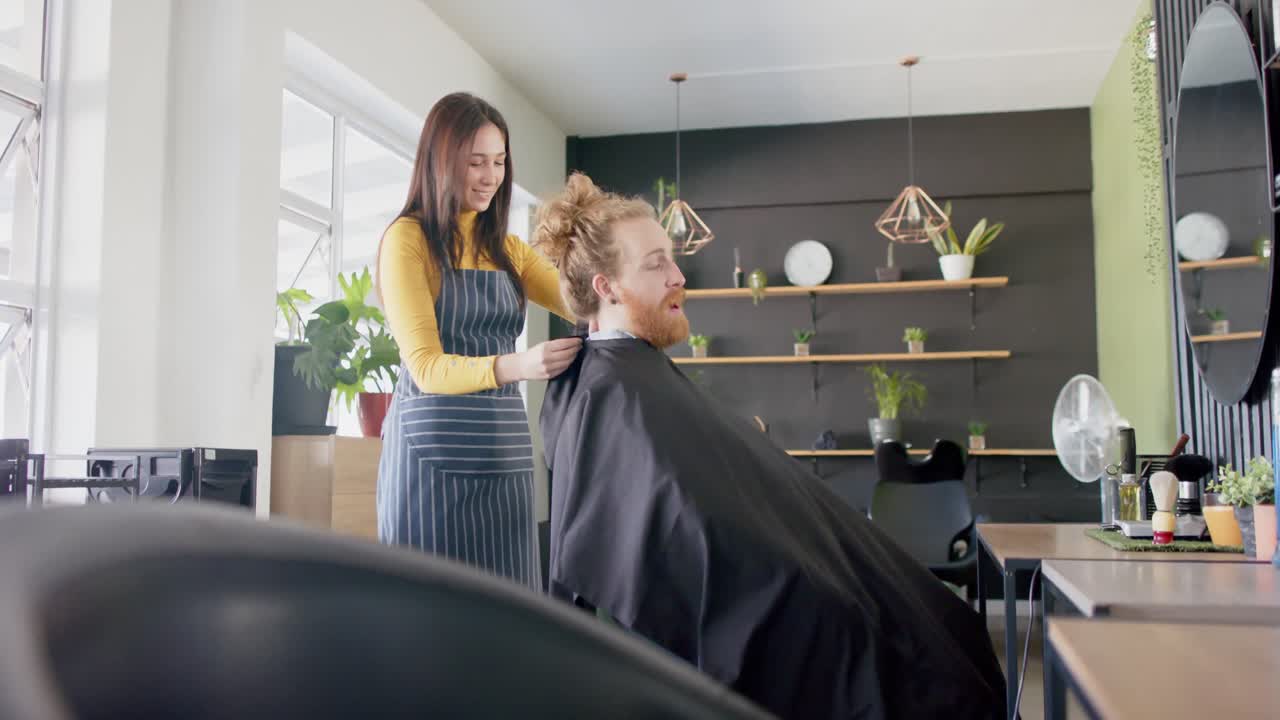 Happy caucasian female hairdresser putting cape on male client at hair salon, in slow motion