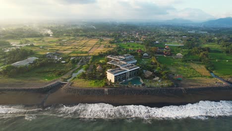 Bird's-Eye-Aerial-Semarapura-Town-Skyline-Balinese-Landscape---Wyndham-Tamansari-Jivva-Resort-Bali-at-Sunset-by-Lepang-Beach,-Farmlands-in-Rural-Countryside-Valley-with-Foggy-Mountains-in-Background