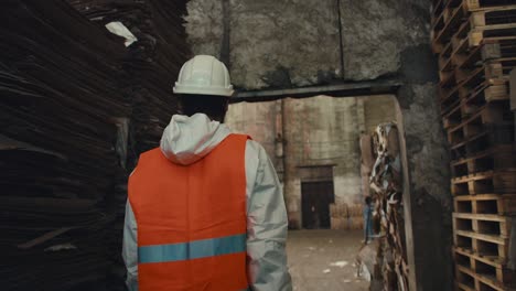 from behind, a brunette man in a white uniform and an orange vest in a white protective helmet walks along the racks with waste paper and goes out into the huge hall of the waste recycling and sorting plant