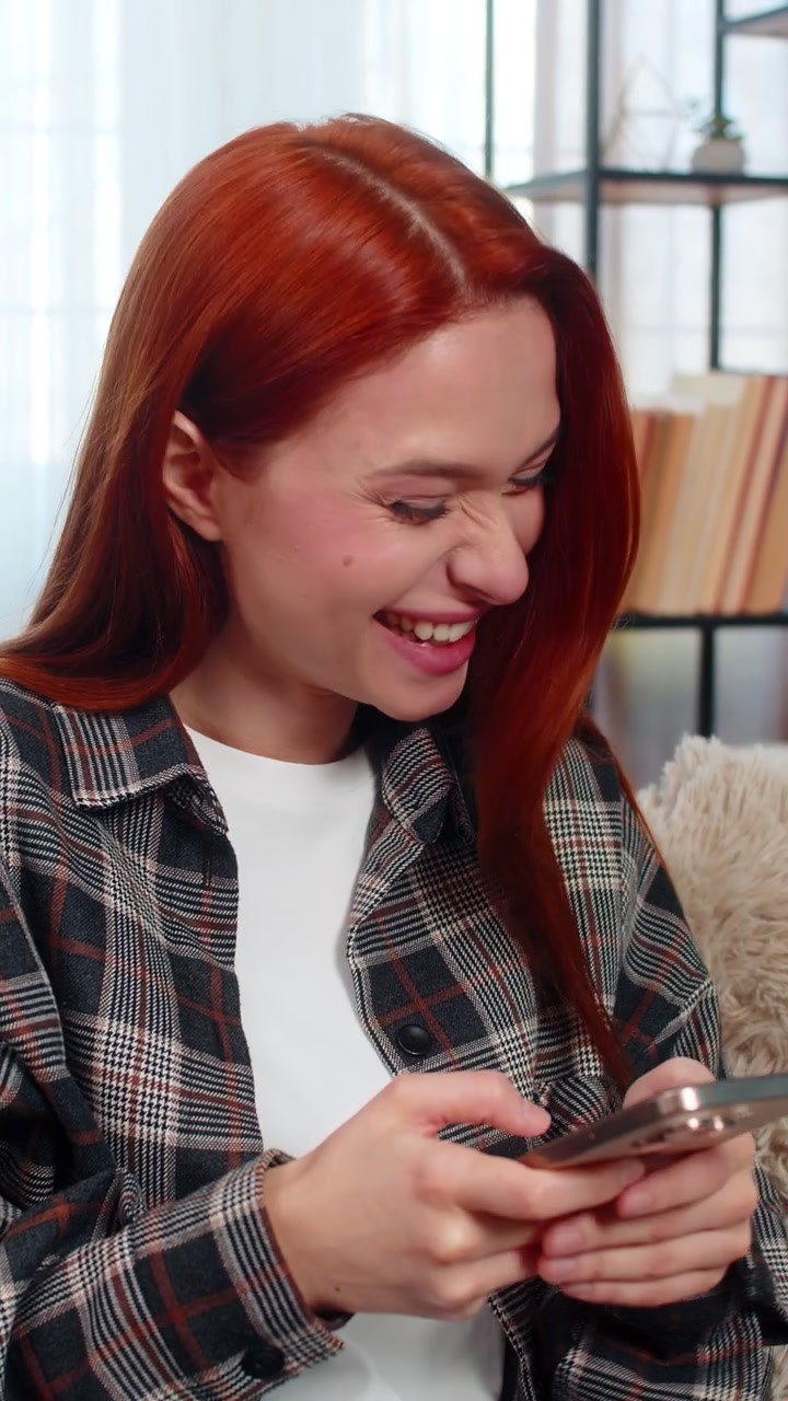 Woman sitting on sofa texting on smartphone with focus and laughing while chatting in messenger app