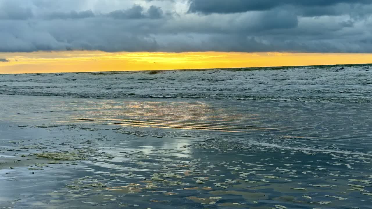 Golden sunset light reflects on wet sand as gentle waves move across the sea, with layered clouds on the horizon creating a calm Mediterranean atmosphere
