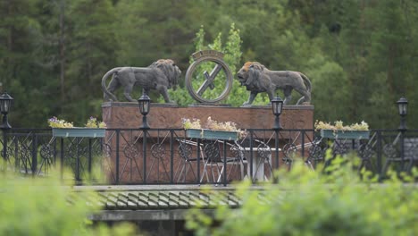 a decorative structure features two lion statues facing each other, with ornate details and greenery surrounding it in the park of the elveseter hotel