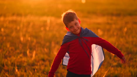 a child in the costume of a superhero in a red cloak runs across the green lawn against the backdrop of a sunset toward the camera.