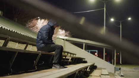 a man wearing a hood is seated alone on stadium bleachers, with a blurred, dimly lit background