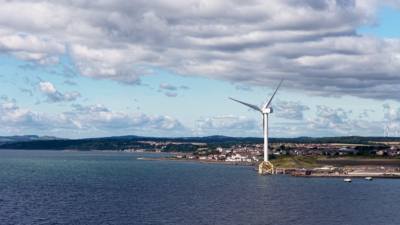 Large wind turbine spins steadily by the coast under partly cloudy daylight, wide static shot